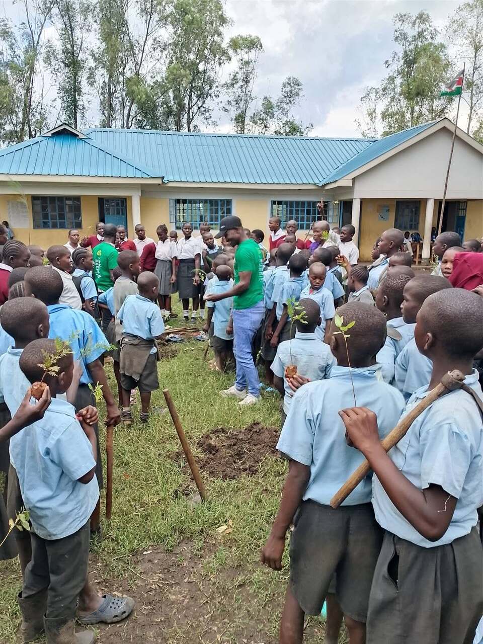 Pupils gathered around a JOFA volunteer during a schoolyard tree-planting session, Kenyan flag flying in the background